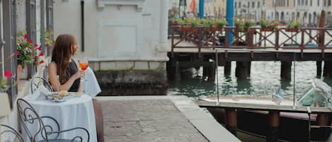A woman sitting at a table with a drink, overlooking a canal with boats and buildings in the background.