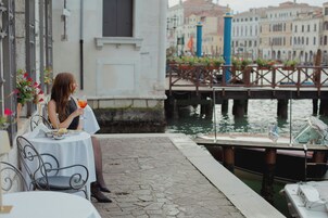 Una mujer sentada a una mesa con una bebida, mirando un canal con barcos y edificios al fondo.