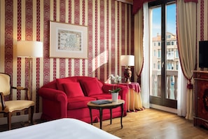 A hotel room with a red sofa, a wooden chair, a small table with a plant, a framed map on the wall, and a balcony with curtains.