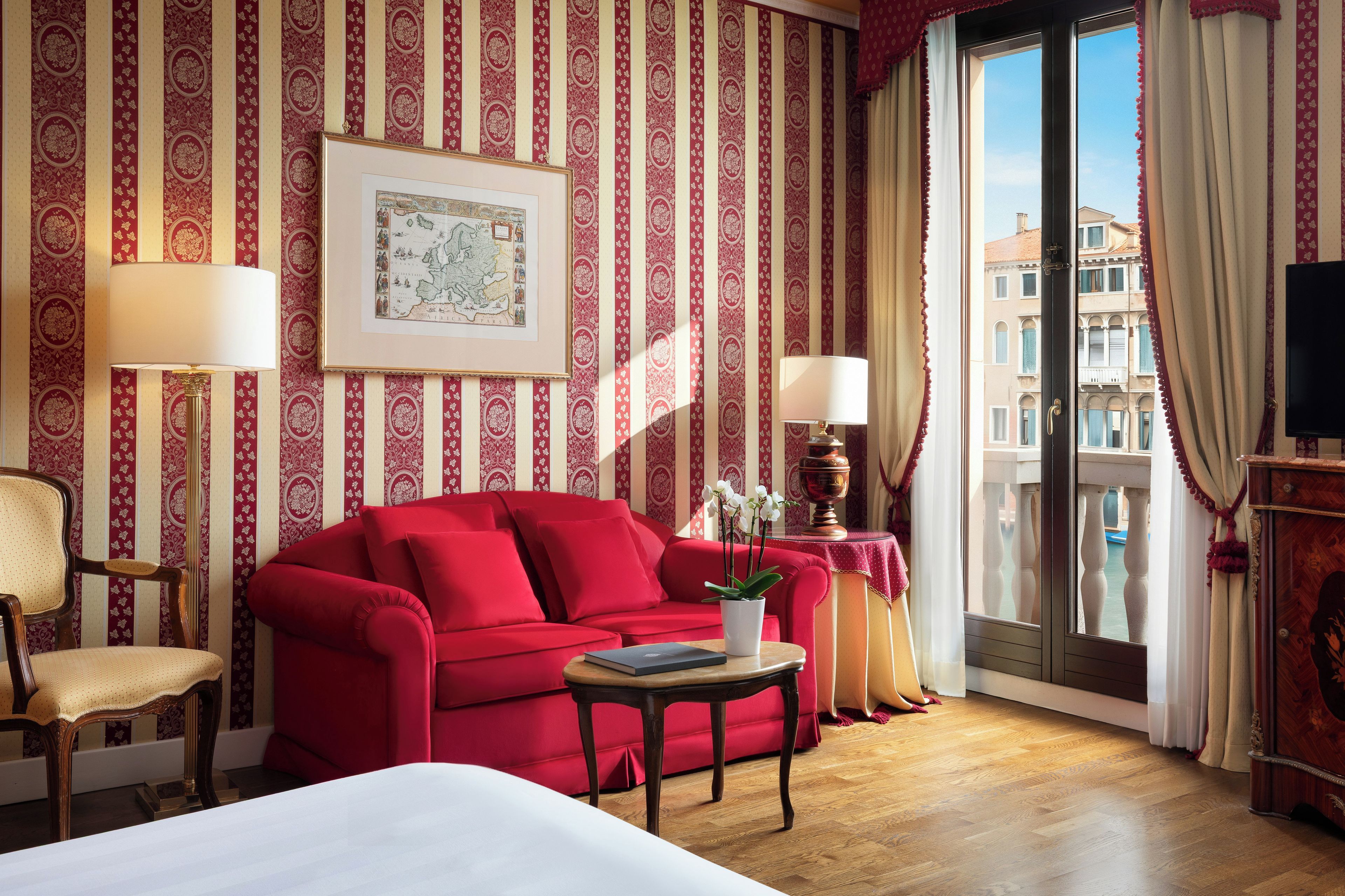 Ein Hotelzimmer mit einem roten Sofa, einem Holzstuhl, einem kleinen Tisch mit Pflanze, einer an der Wand hängenden Karte im Bilderrahmen und einem Balkon mit Vorhängen.