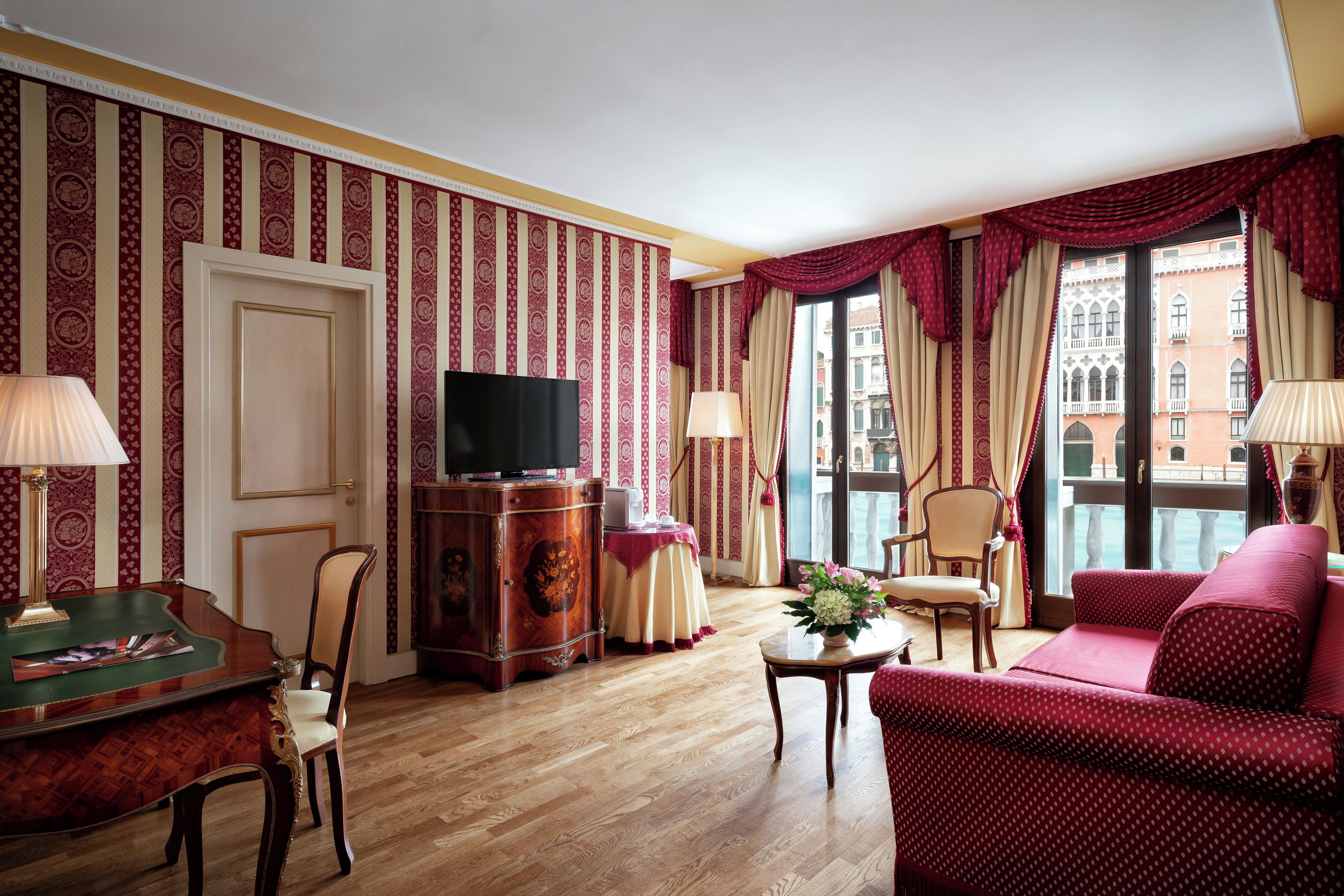 Habitación de hotel con piso de madera, televisor, mesa de comedor con sillas, sofá y ventana con cortinas.