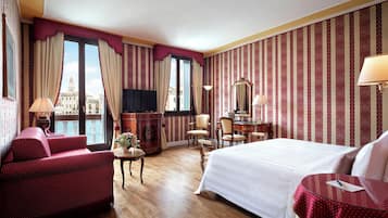 Quarto de hotel com cama, escrivaninha, cadeira, televisão e vista para um prédio pela janela.