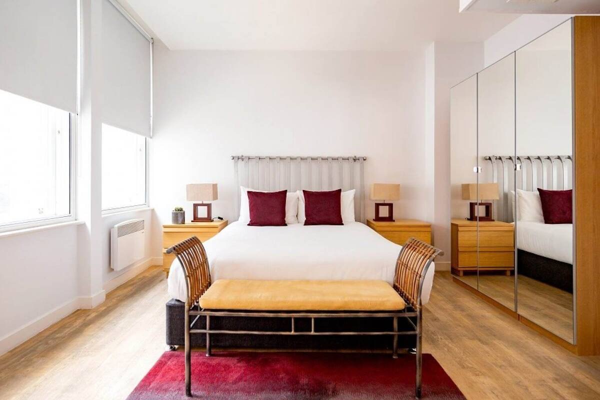 A modern hotel room with a large bed, a wooden bench, a mirror, and a window.