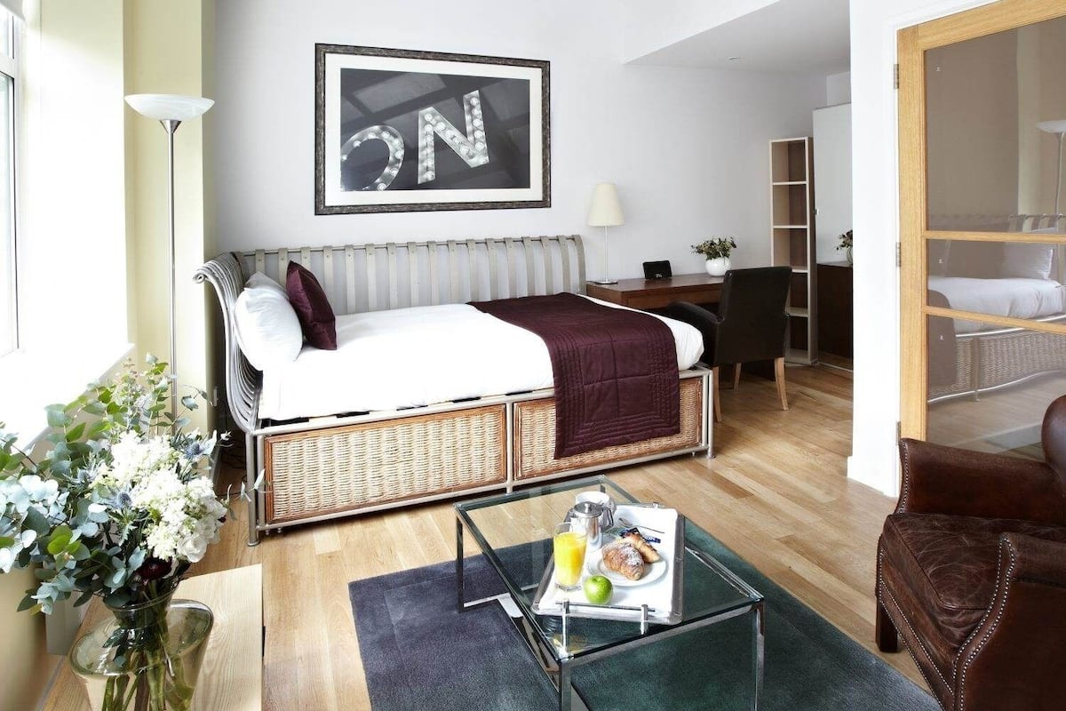 A compact room with a bed, a small table with breakfast items, a chair, a lamp, and a framed 'ON' sign on the wall.