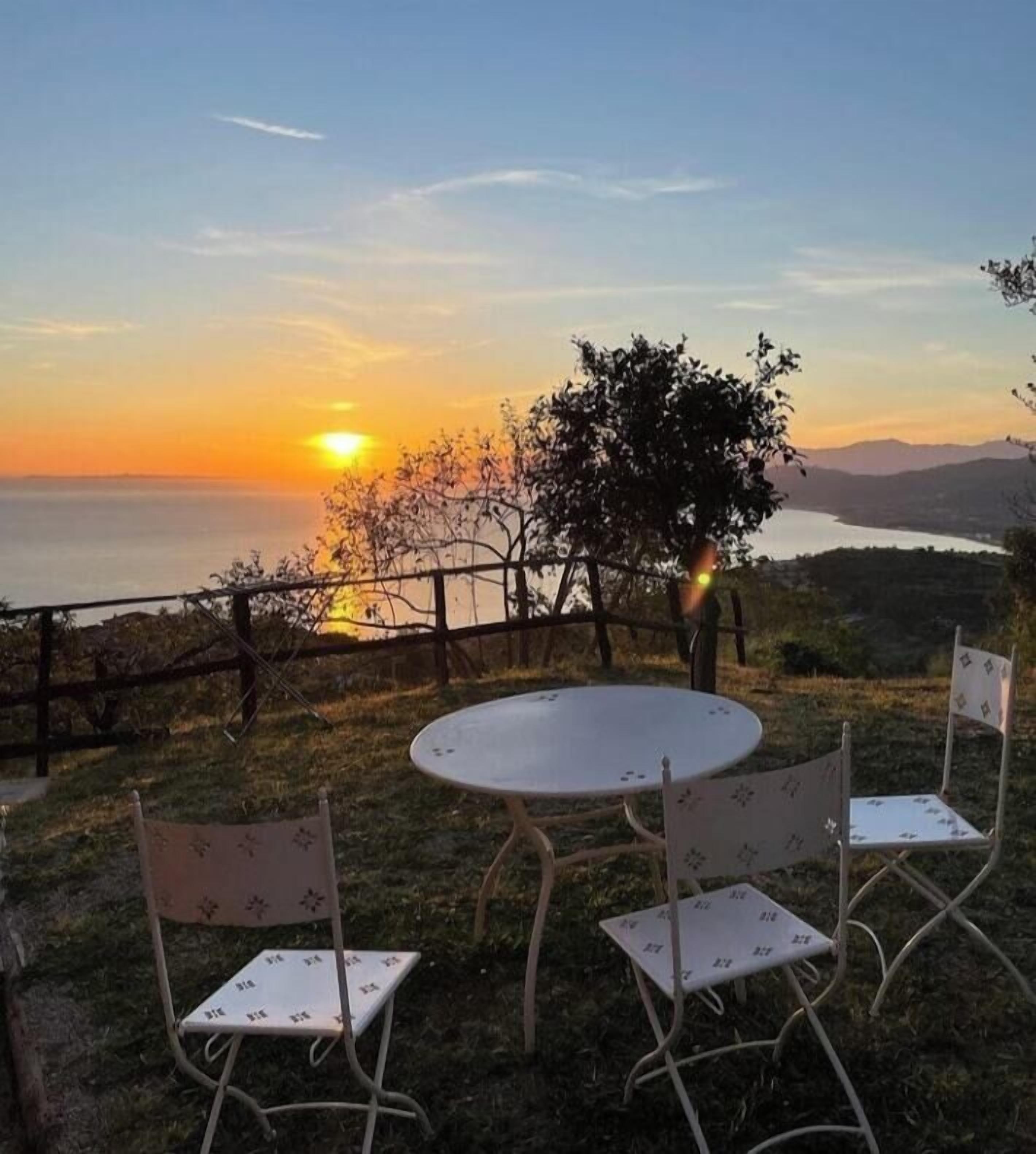 A round table with two chairs, a view of a sunset over a body of water, and a tree in the foreground.