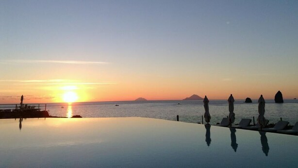 Una piscina infinita con vistas a la puesta de sol sobre el océano, sillas de descanso y sombrillas.