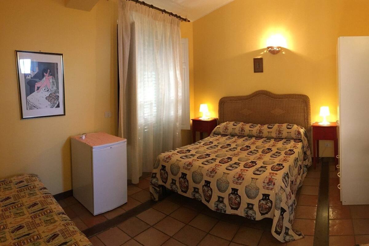 Ein kleines Hotelzimmer mit einem Bett, einem Kühlschrank, einem Nachttisch mit Lampe, einem Bild an der Wand und einer Tür rechts.