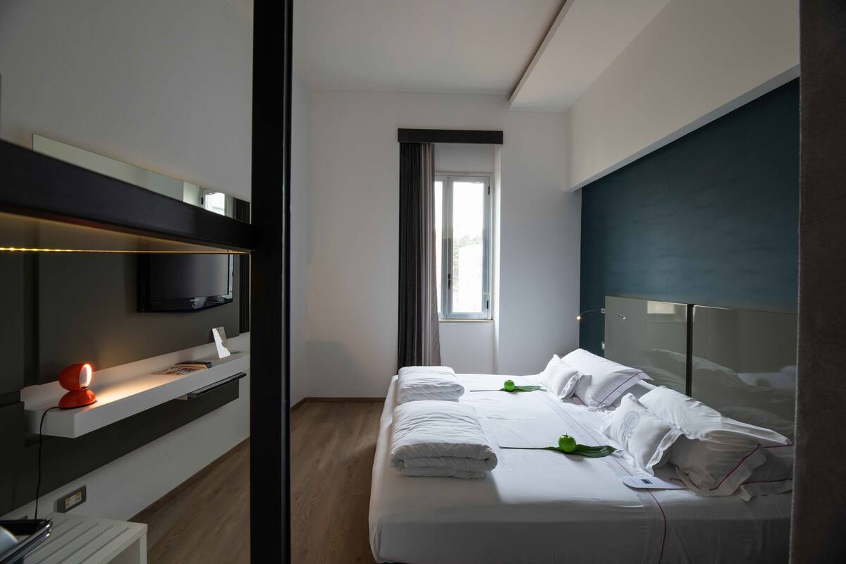 Una camera d'albergo moderna con un letto, una TV, una scrivania con lampada e una finestra.