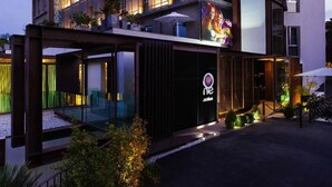 A modern hotel with a glass facade, outdoor seating area, and a visible logo on the building. - UNA Hotels One Siracusa Wellness & SPA (Syracuse)