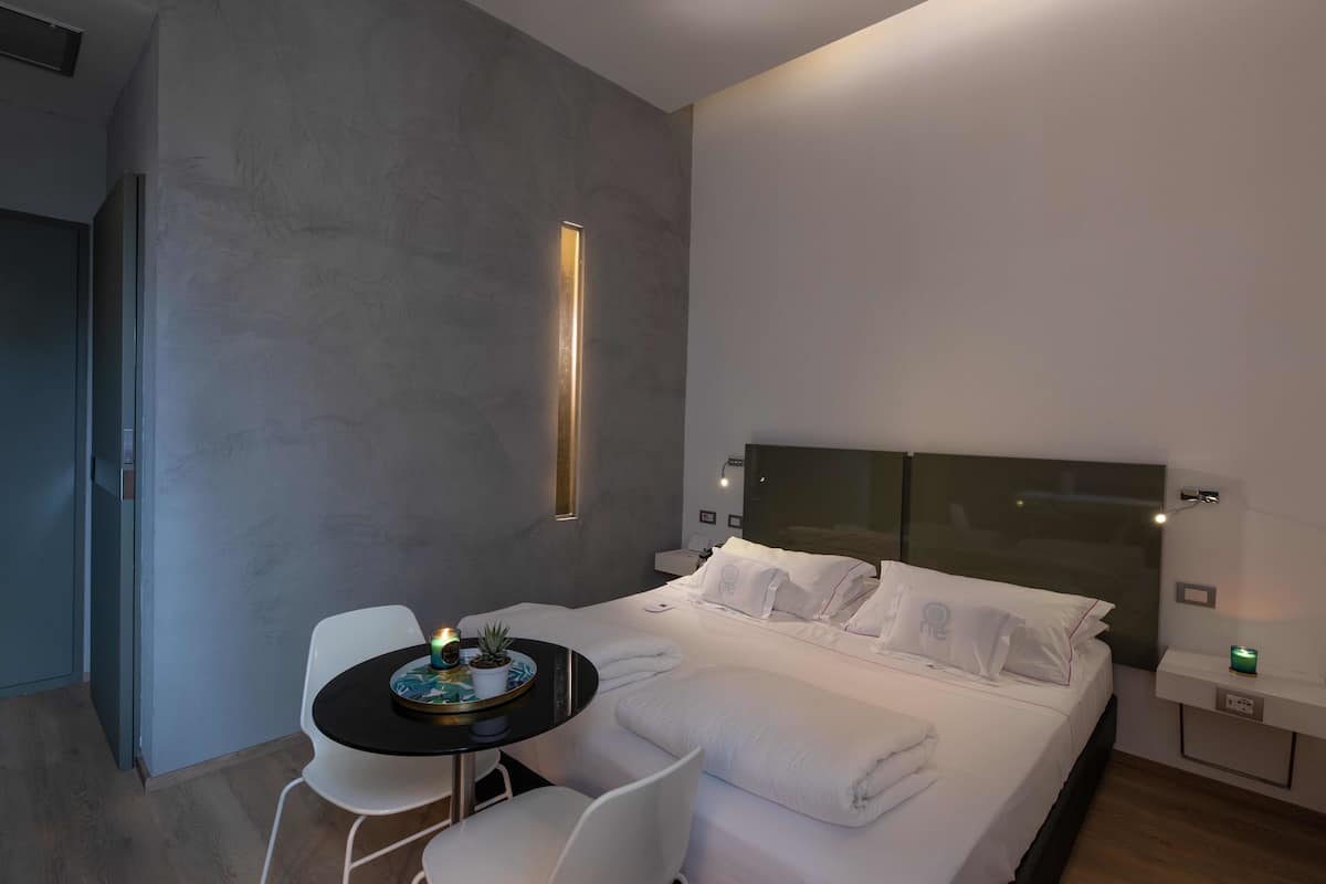 Una camera d'albergo moderna con un letto grande, un comodino con una lampada, un tavolino rotondo con una pianta e una sedia.