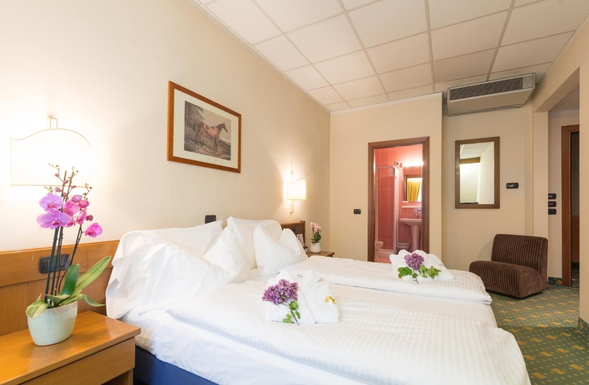 Ein Hotelzimmer mit zwei Betten, einem Nachttisch mit Lampe, einem gerahmten Bild an der Wand und einem durch eine offene Tür einsehbaren Badezimmer.