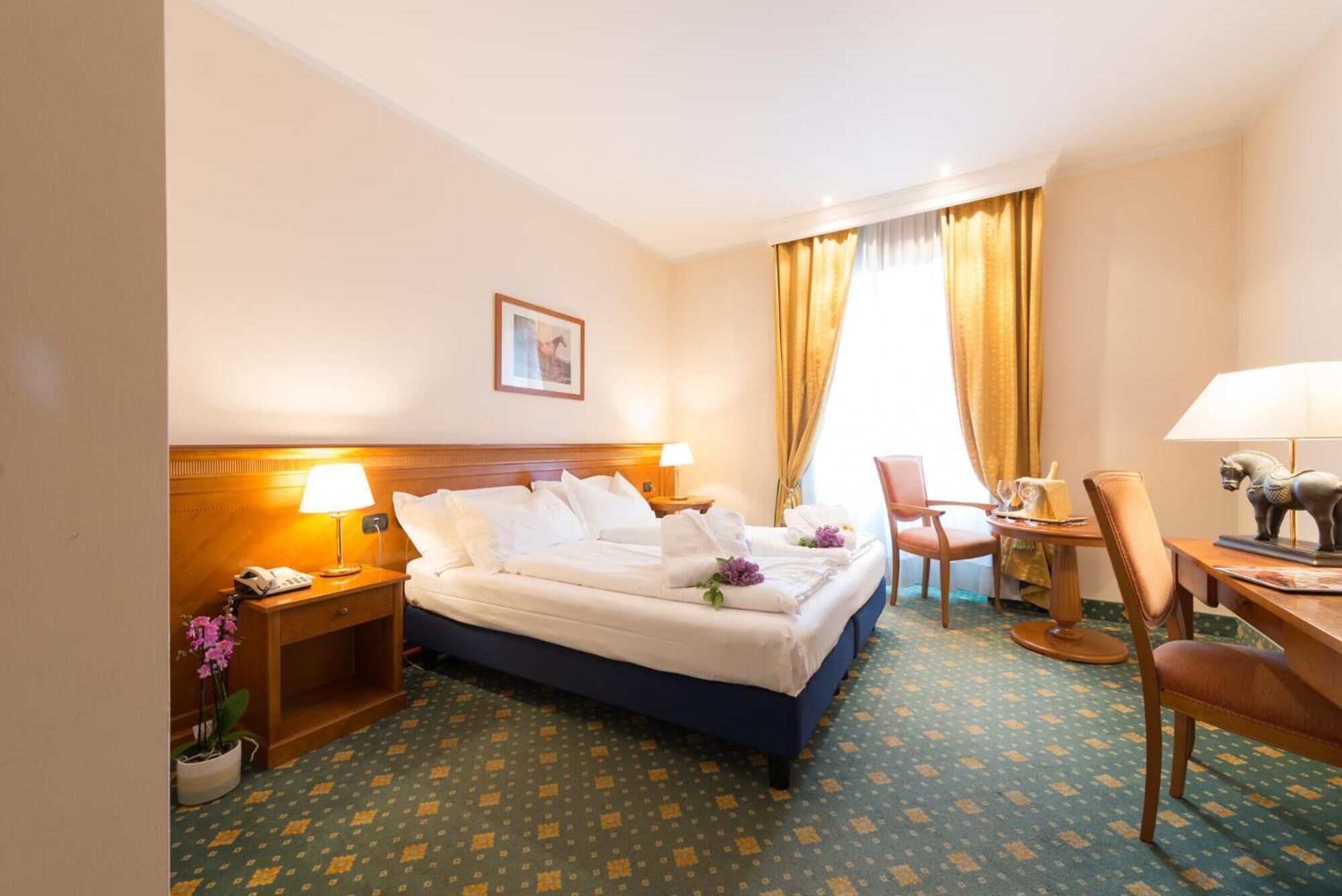Una camera d'albergo con un letto, comodini, una scrivania con una sedia, una lampada e una finestra con tende.