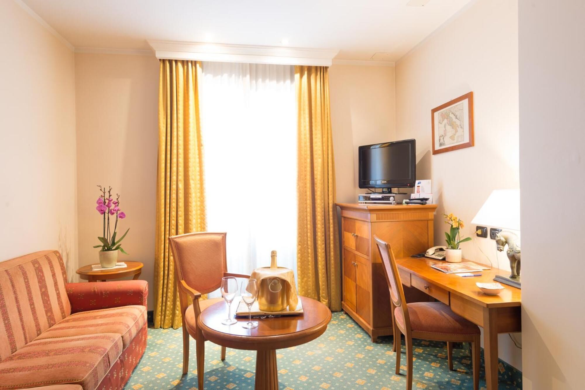 Una habitación de hotel con sofá, una pequeña mesa redonda con tetera y vasos, un escritorio de madera con computadora y teléfono, un televisor y una ventana con cortinas.