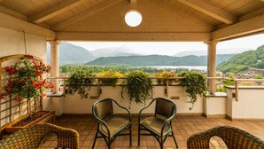 A covered balcony with two wicker chairs, a potted plant with red flowers, and a view of a lake and mountains. - Hotel Eden Levico Terme (Levico Terme)