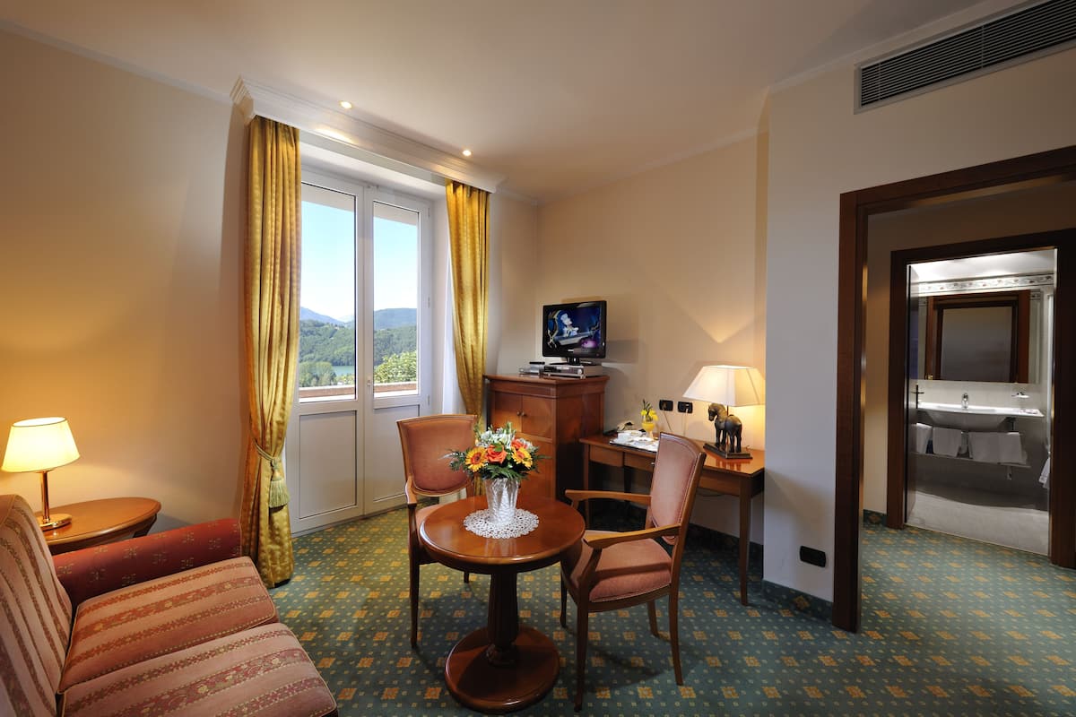 Ein Hotelzimmer mit einem Bett, einem Fernseher, einem Schreibtisch mit Lampe, einem Sessel, einem kleinen Tisch mit Blumenvase und Blick ins Freie durch ein Fenster.