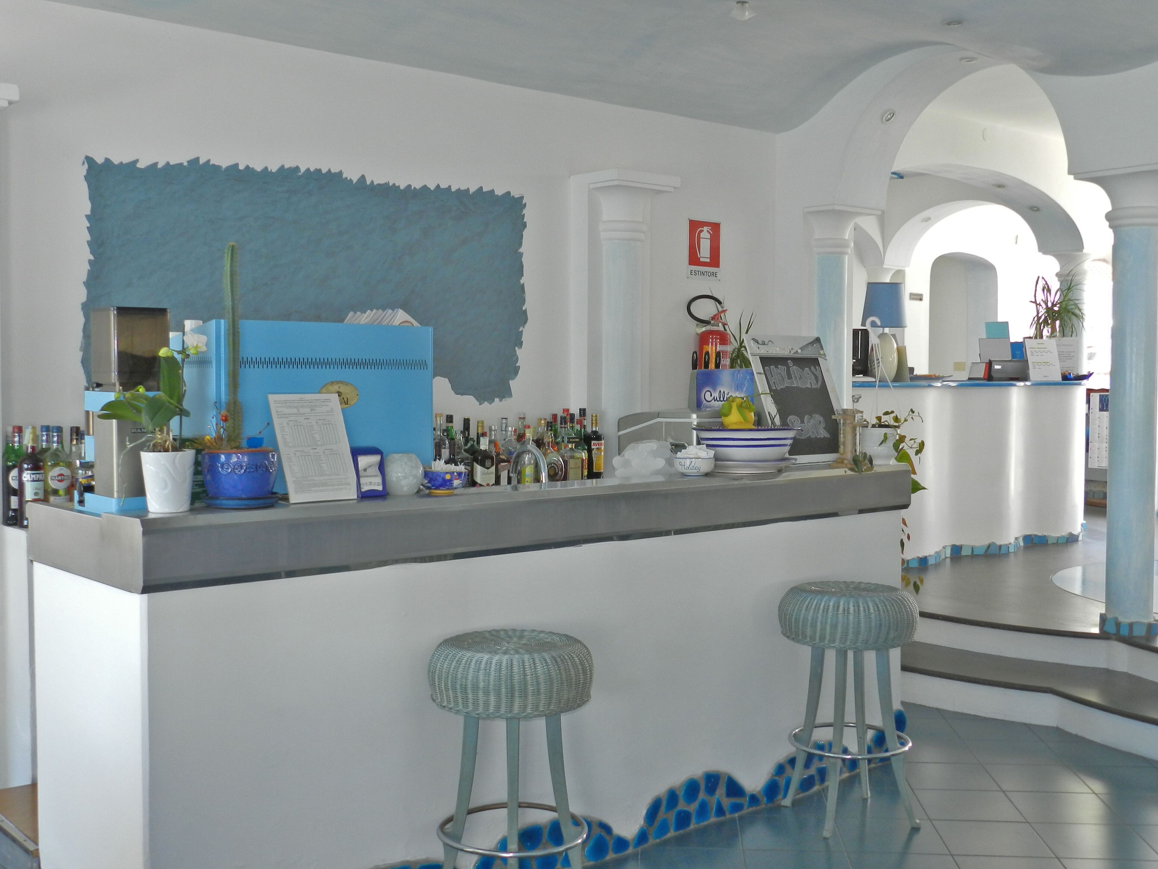 Zona de bar con taburetes, una barra con diversas botellas, una pared decorativa azul y un letrero que dice "Holiday Bar".