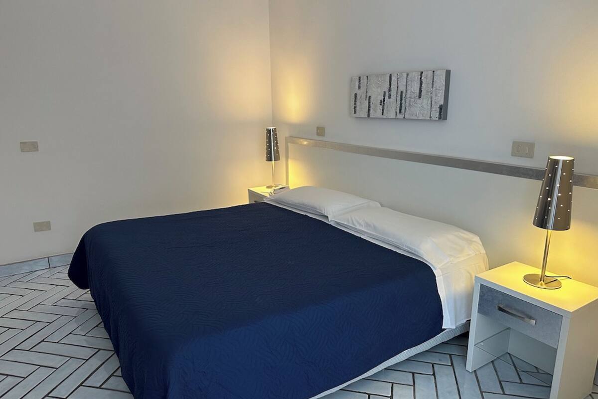 A neatly made bed with a dark blue bedspread and white pillows, flanked by two bedside tables with lamps, one of which has a metallic base.