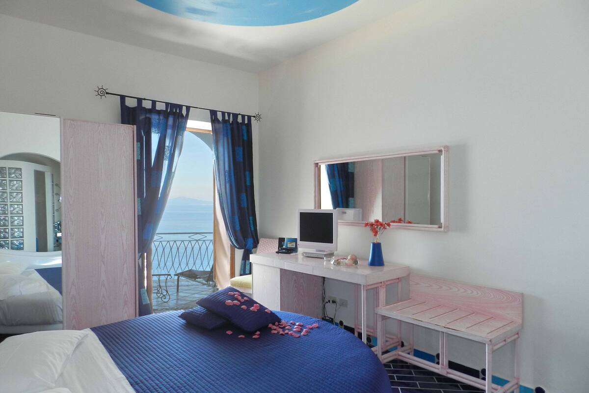 A bedroom with a bed, a desk, a chair, a mirror, and a view of the sea.