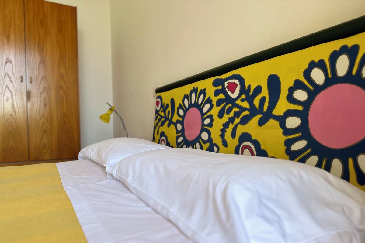 A bed with white bedding and a yellow blanket, a wooden wardrobe, and a wall with a colorful floral pattern.