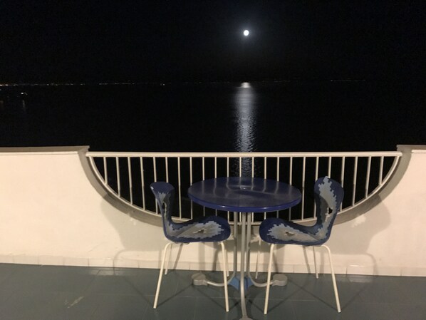 A balcony with a table and two chairs, overlooking a body of water at night.