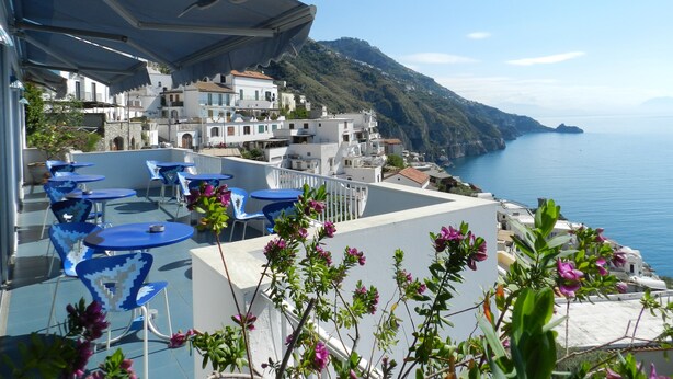 Una terraza con mesas y sillas azules, con vistas a un pueblo costero con edificios y vista al mar.