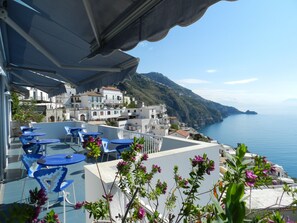 Una terraza con mesas y sillas azules, con vistas a un pueblo costero con edificios y vista al mar.