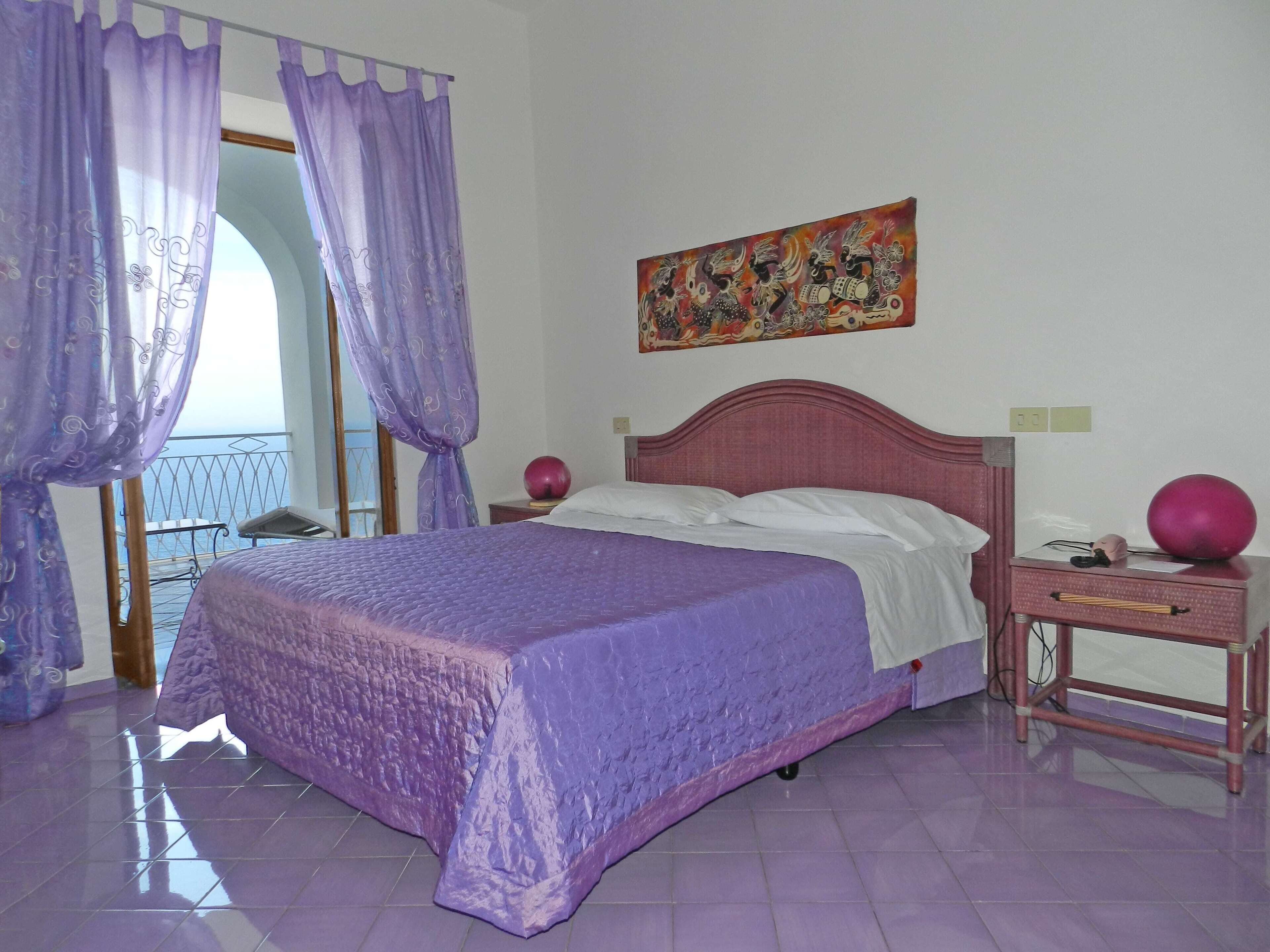 Un dormitorio con cama, cortinas moradas, un cuadro y vistas al balcón.