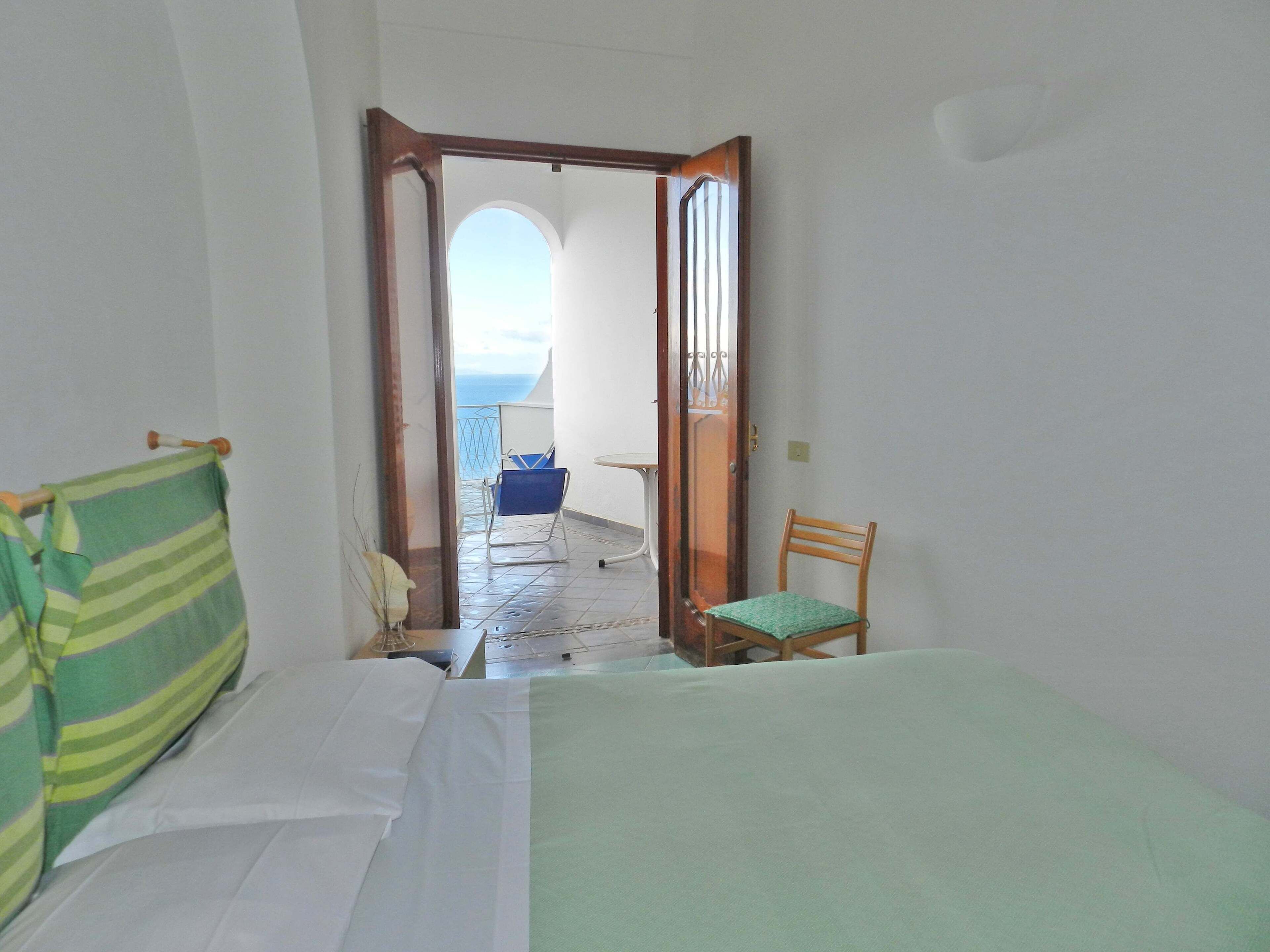 A room with a bed, a chair, and a view through an open door to a balcony with a chair and a table.