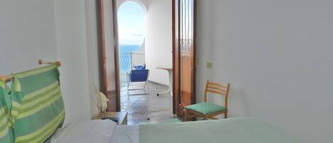 A room with a bed, a chair, and a view through an open door to a balcony with a chair and a table.
