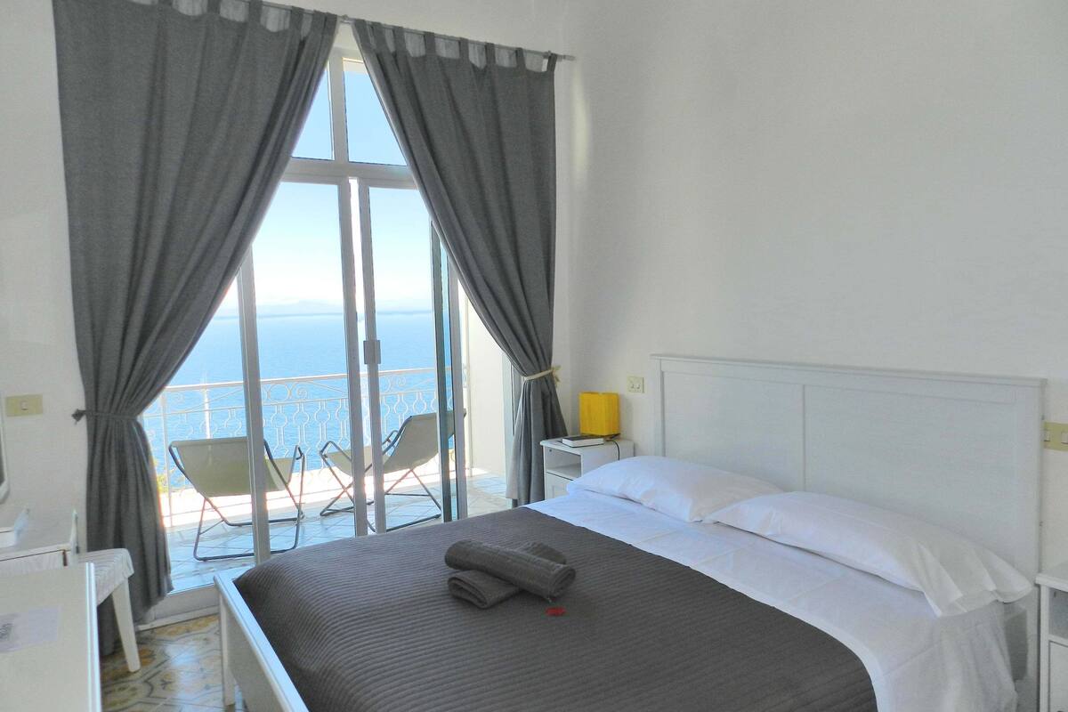 A hotel room with a bed, a bedside table with a lamp, a chair, and a view of the sea through large windows.