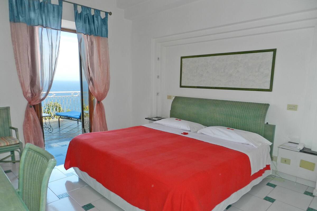 A hotel room with a bed, a chair, a table, and a view of the sea through a balcony.