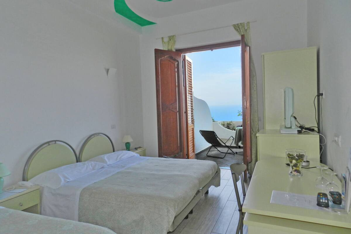 A hotel room with two beds, a desk with a mirror, and a view of the sea through an open door.