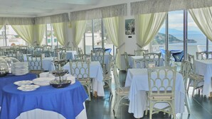 Un restaurante con varias mesas y sillas, ventanas amplias con cortinas, y vista al mar.