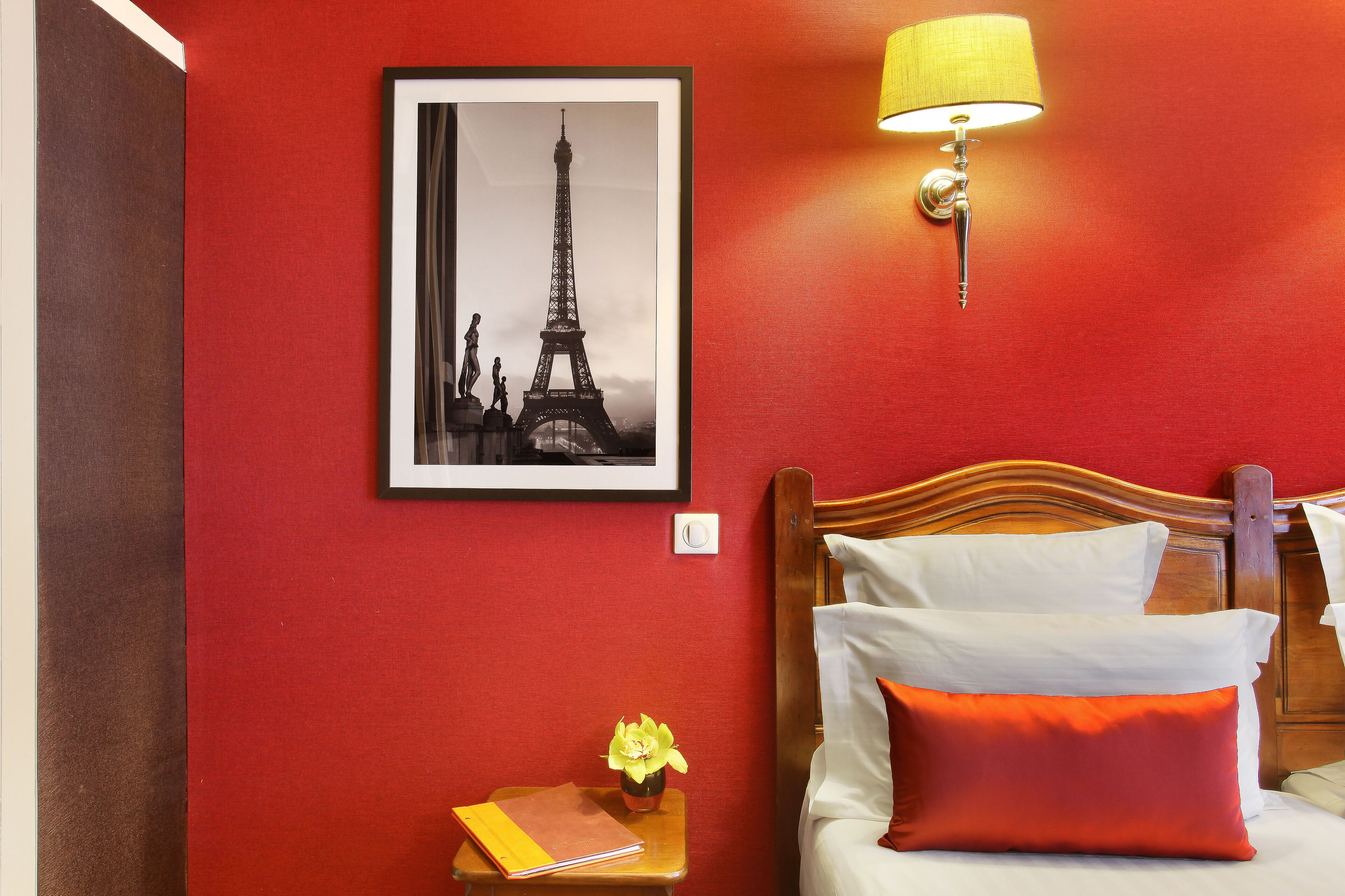 A hotel room with a red wall, a framed picture of the Eiffel Tower, a bedside lamp, a wooden headboard, white and orange pillows, a small table with a vase of flowers, and a nightstand with a book and a lamp.