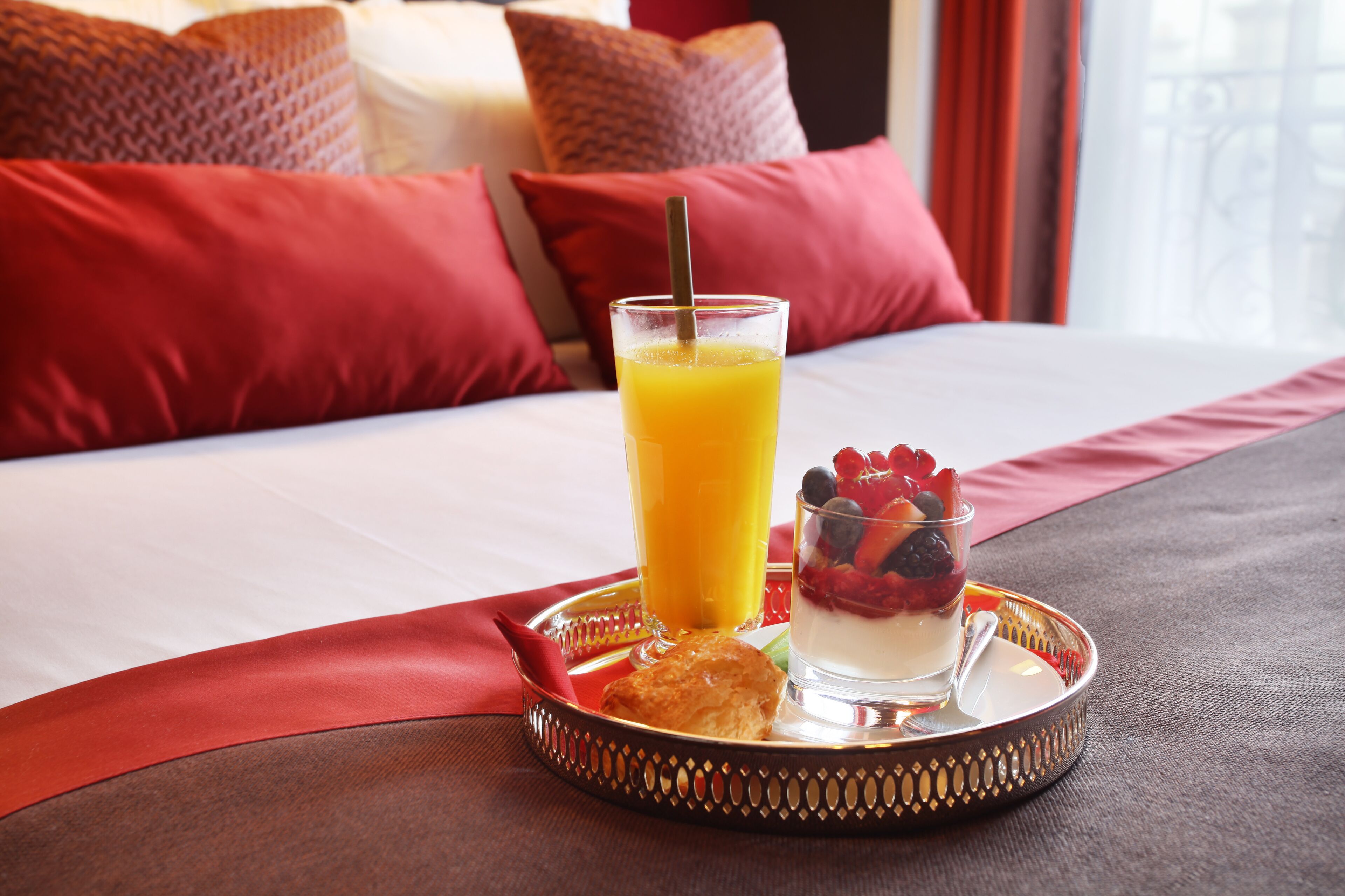 Bandeja de desayuno con jugo de naranja, yogur con bayas y un pastel sobre una cama con almohadillas y cubrecama rojos.