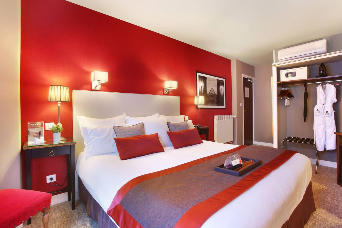 Ein Hotelzimmer mit einem großen Bett, roten Wänden, einem Nachttisch mit Lampe, einem kleinen Tisch mit Tablett und einem Kleiderhaken mit Bademänteln.