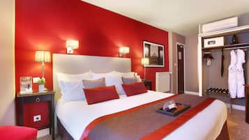 Habitación de hotel con una cama grande, paredes rojas, una mesita de noche con lámpara, una pequeña mesa con bandeja y un perchero con batas.