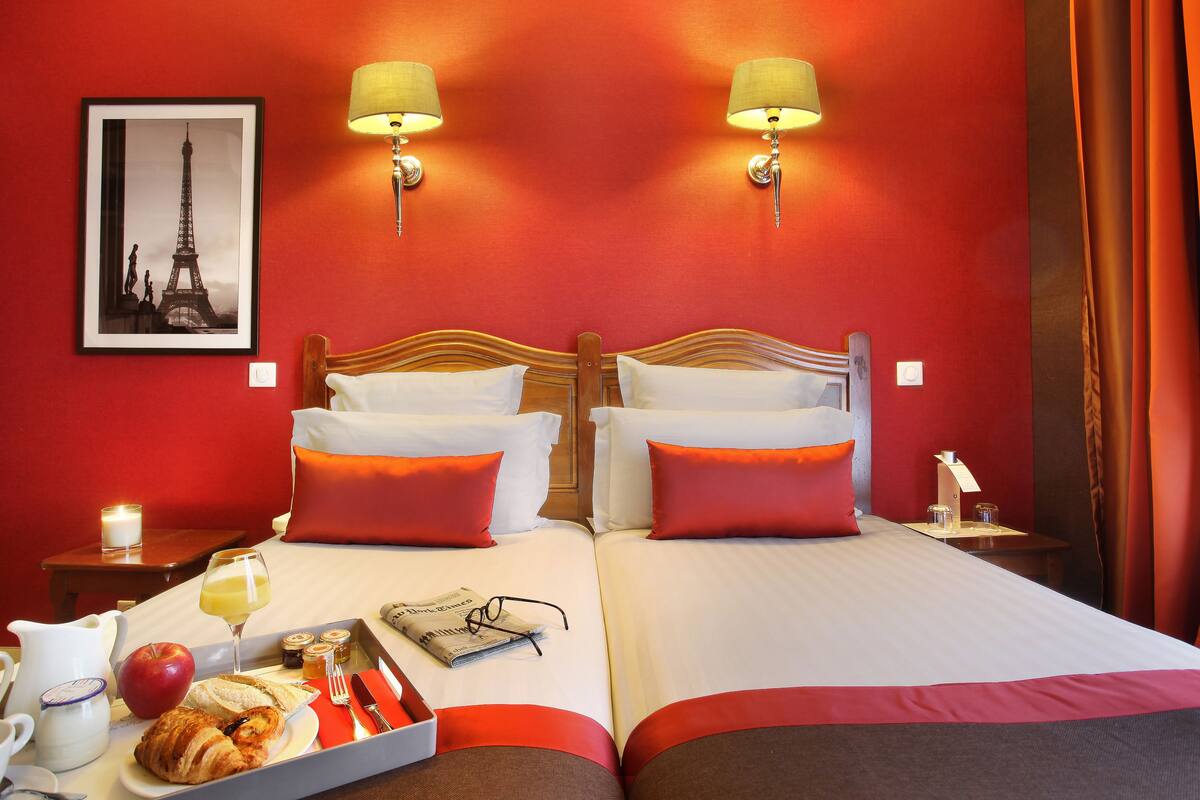 Ein Hotelzimmer mit zwei Betten, roter und weißer Bettwäsche, einem Frühstückswagen mit Croissants, einem Apfel und einem Glas Saft. An der Wand hängt ein gerahmtes Bild vom Eiffelturm.
