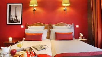 A hotel room with two beds, red and white bedding, a breakfast tray with croissants, an apple, and a glass of juice. There is a framed picture of the Eiffel Tower on the wall.