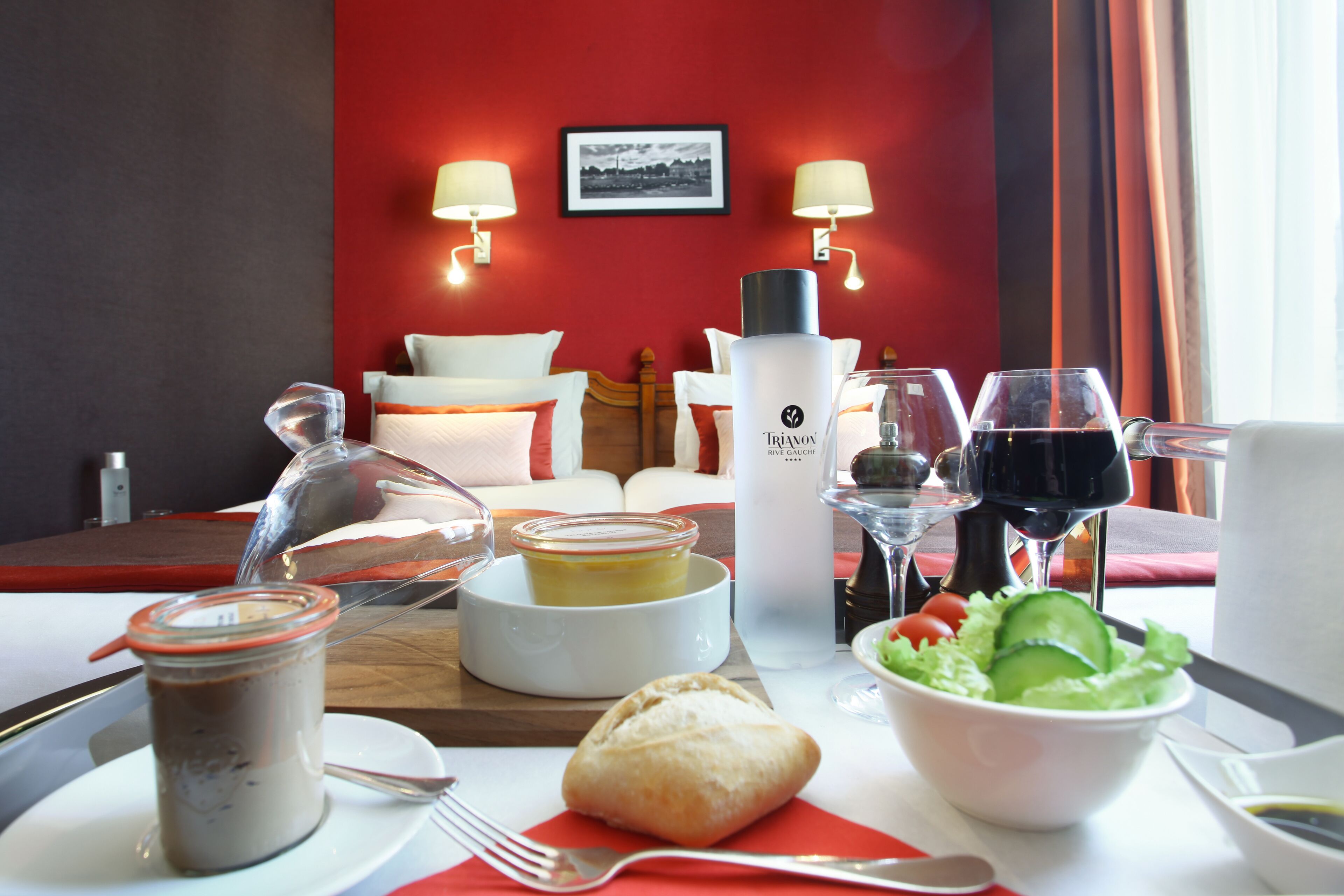 A hotel room with a dining table set with food, a bottle of Trionox, wine glasses, and a bed with pillows and a headboard.