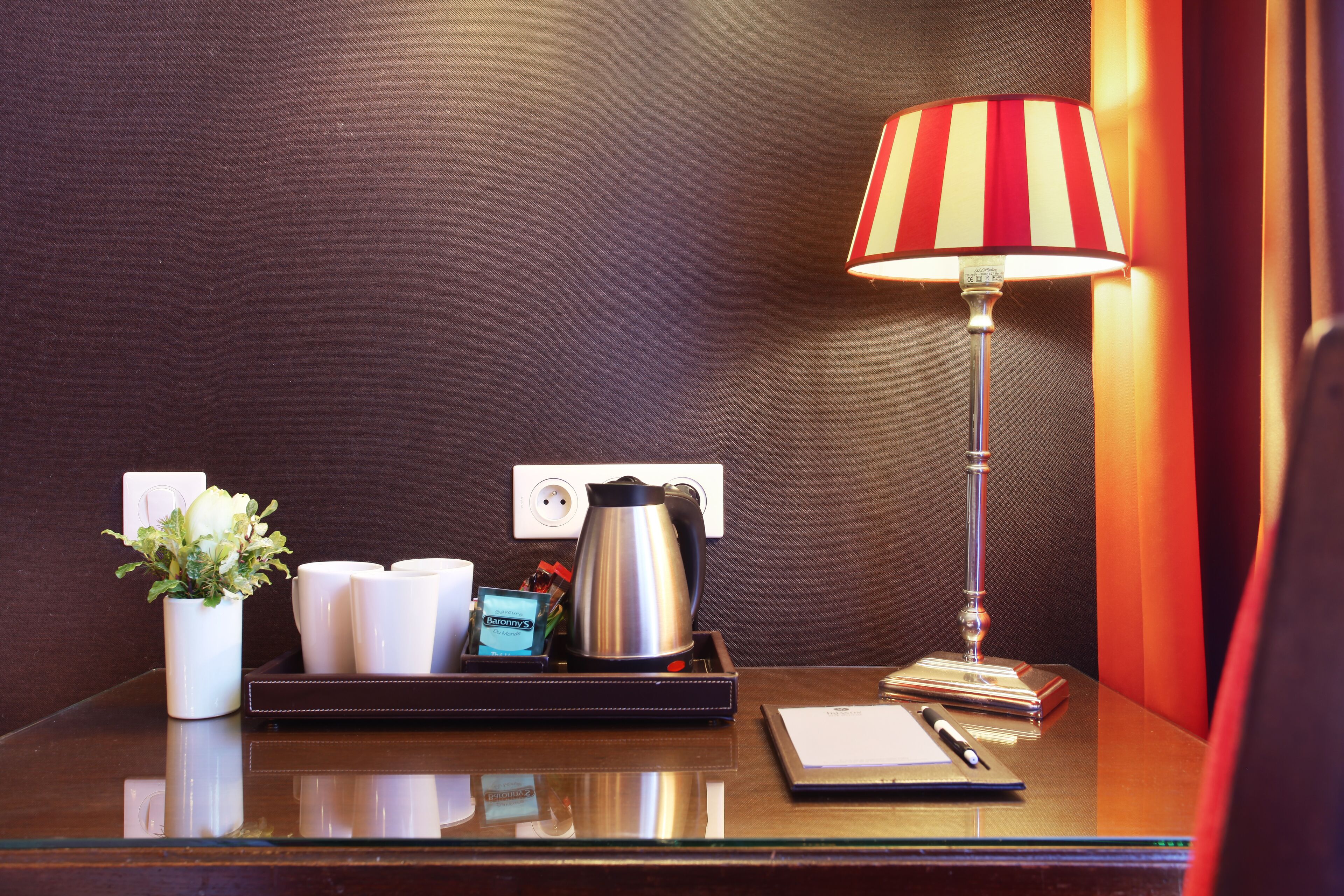 Un escritorio de hotel con una lámpara, una cafetera, una bandeja con tazas y una planta, además de un bloc de notas.