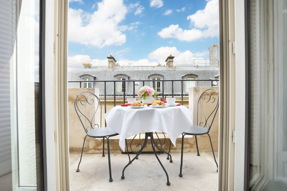Ein Balkon mit Tisch für zwei Personen, einer Blumenvase und Blick auf Gebäude im Hintergrund.