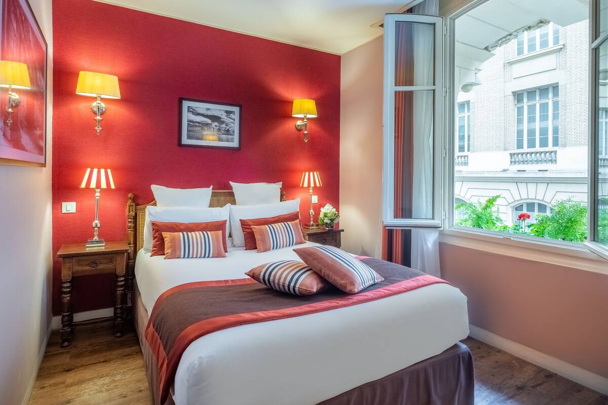 Ein Hotelzimmer mit einem großen Bett, zwei Nachttischen, einem Fenster mit Blick auf Gebäude und roten Wänden.