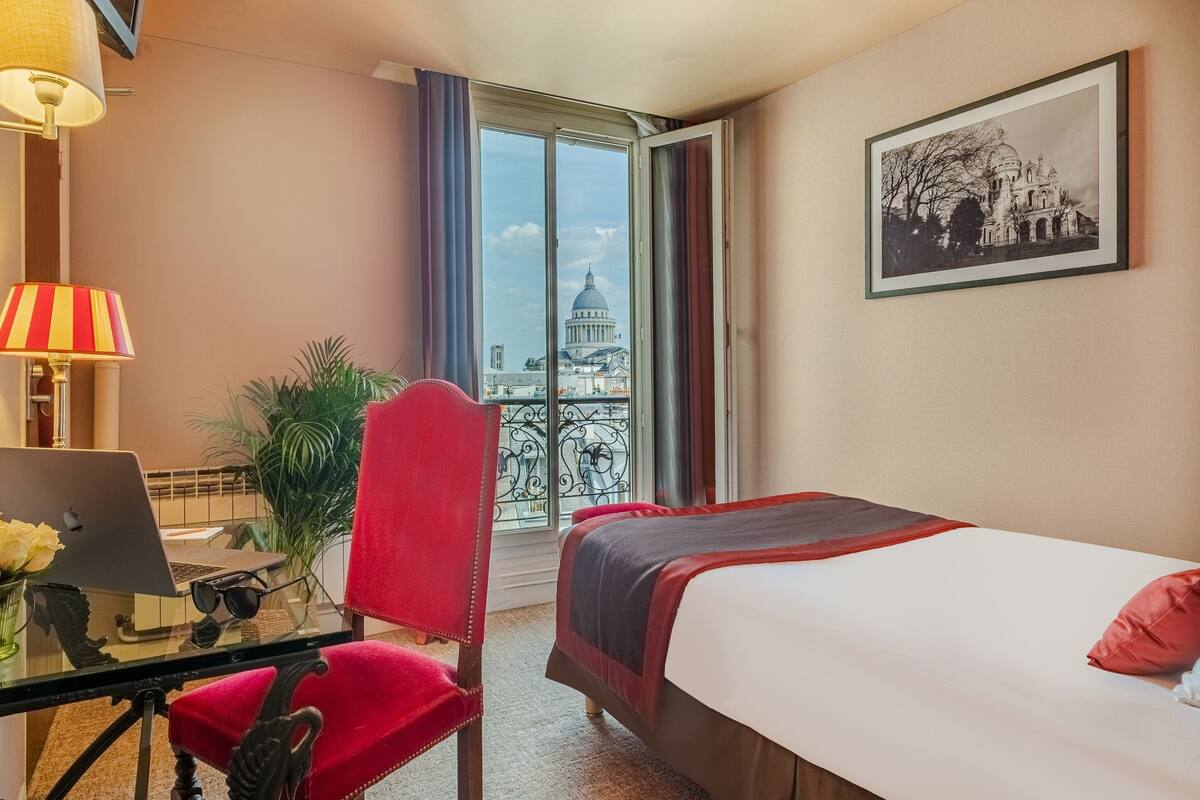 Ein Hotelzimmer mit Bett, Schreibtisch mit Laptop, Stuhl, Lampe und Blick auf ein Gebäude durch das Fenster.