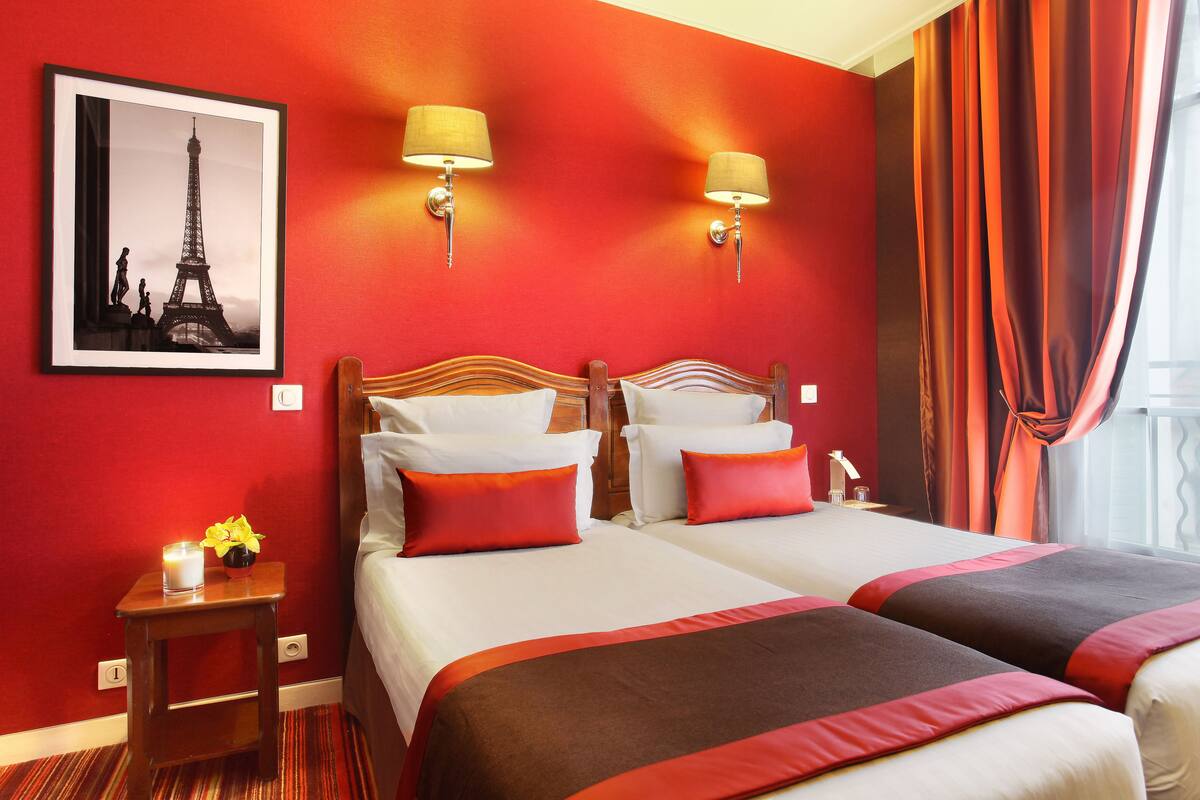 Ein Hotelzimmer mit zwei Betten, roten Wänden, einem gerahmten Bild des Eiffelturms und zwei Wandleuchten.