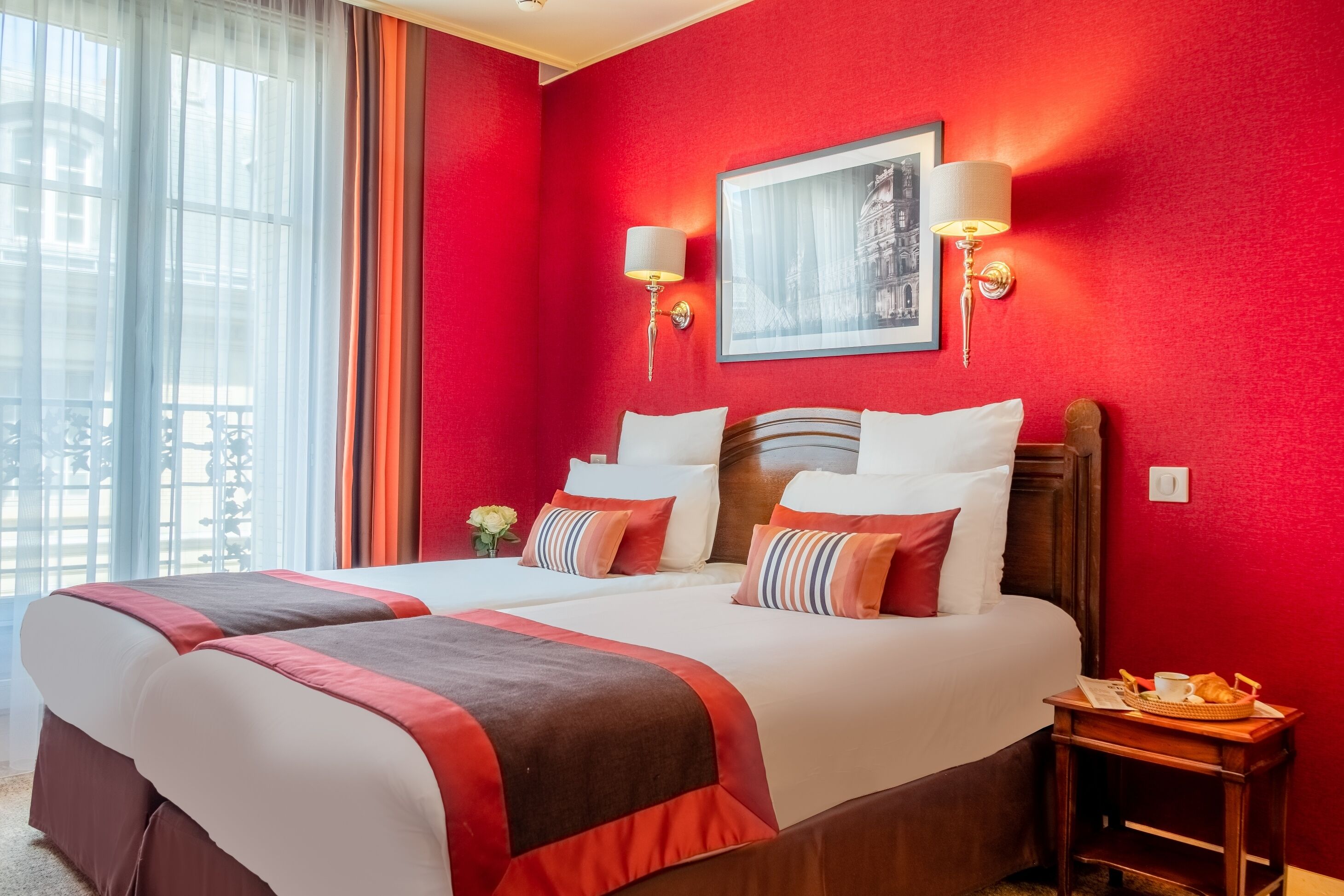 A hotel room with a large bed, two bedside tables, a red wall, and a window with curtains.