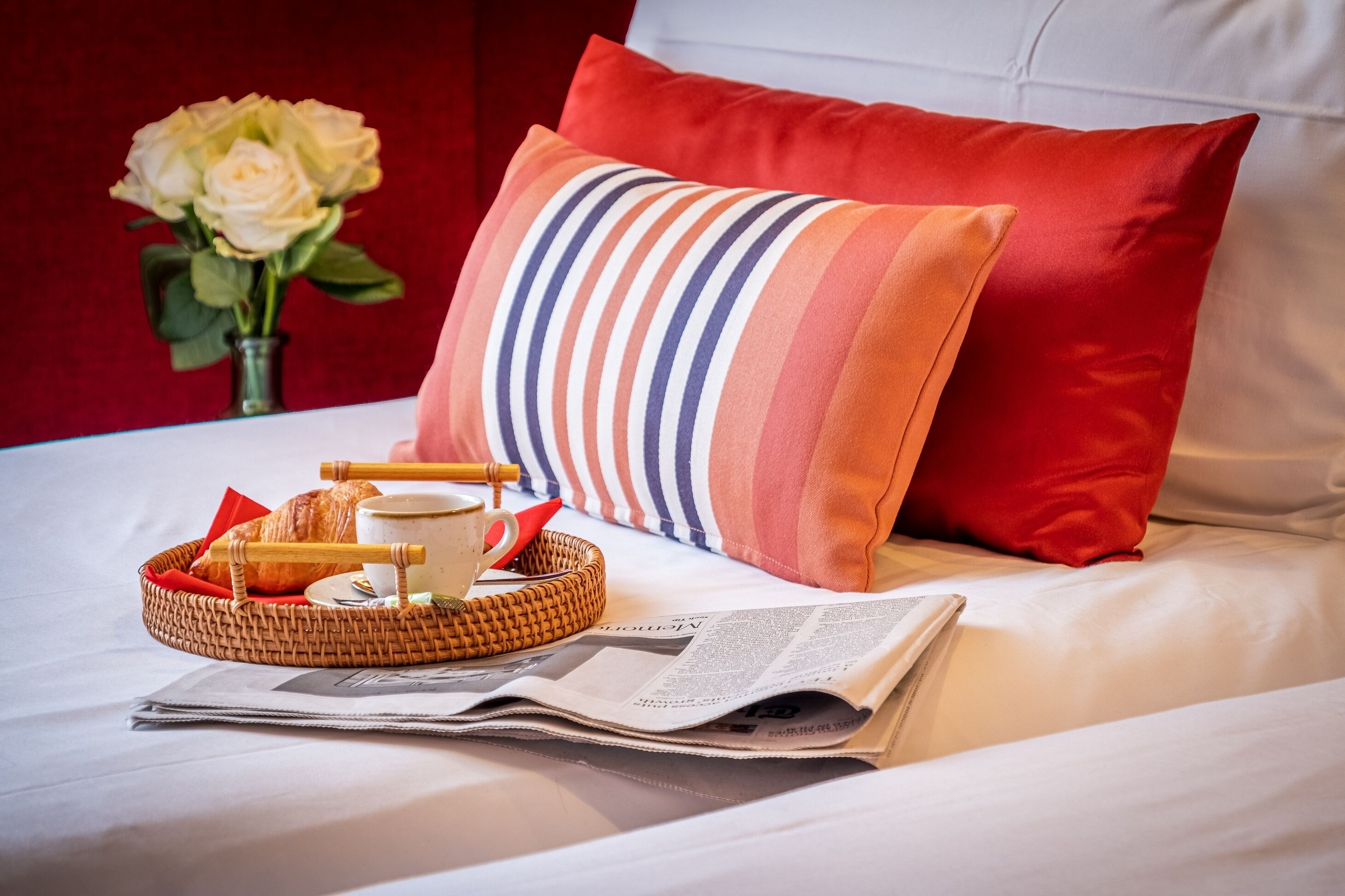 A neatly made bed with a striped pillow, a basket with croissants, a cup, and a newspaper.