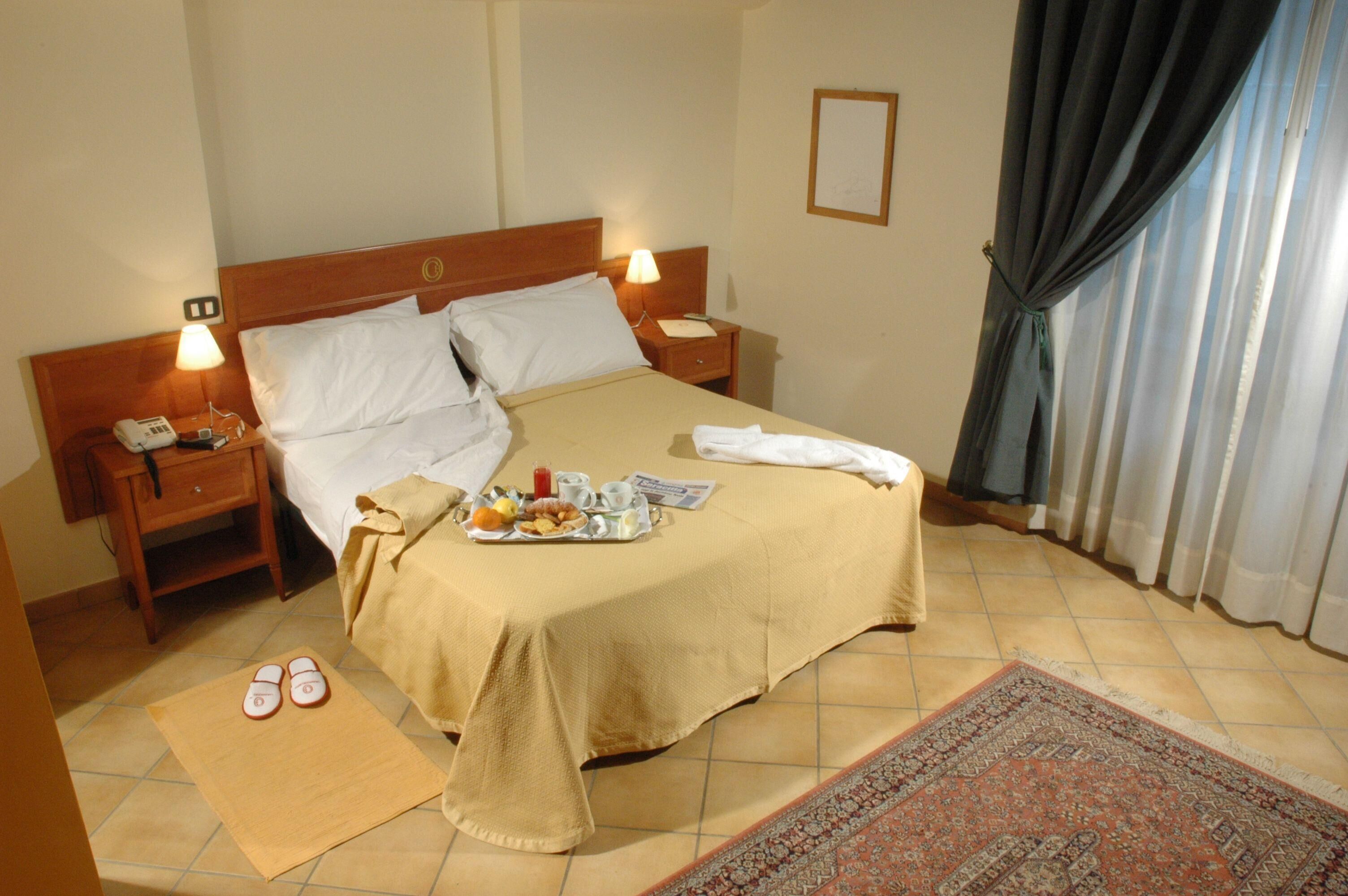 Habitación de hotel con cama, dos mesitas de noche, un atril, un par de zapatillas, una bandeja con alimentos para el desayuno y una alfombra estampada.
