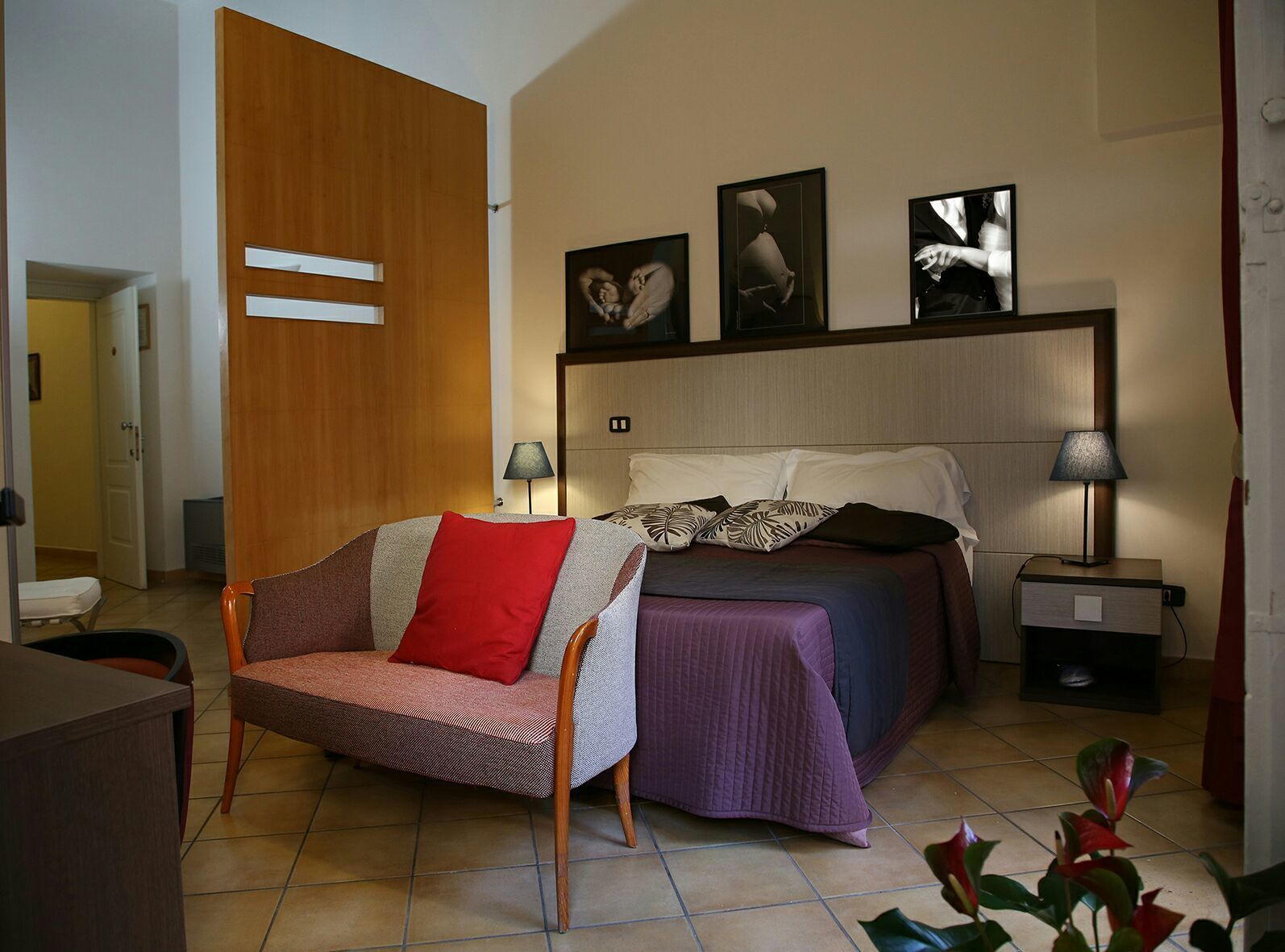 A bedroom with a bed, a chair, a bedside table, a lamp, and wall art.