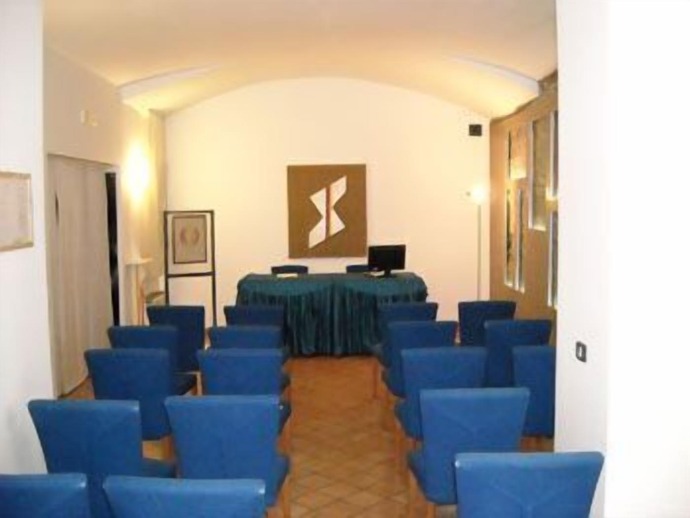 Una sala conferenze con file di sedie blu, un tavolo con tovaglia scura e un'ampia opera d'arte astratta sulla parete.
